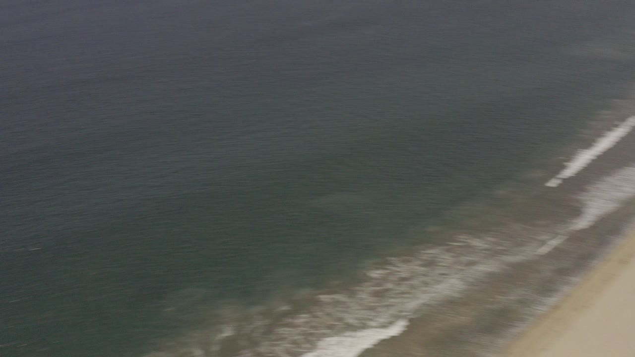 Aerial view in Los Cabos, Mexico showing dark blue ocean and white surf approaching a pale sandy beach, with no visible people at this moment.