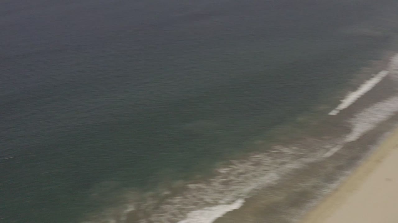 Aerial Los Cabos beach view with white waves rolling toward a broad sand shoreline and the edge of resort development beginning to appear at the upper right.