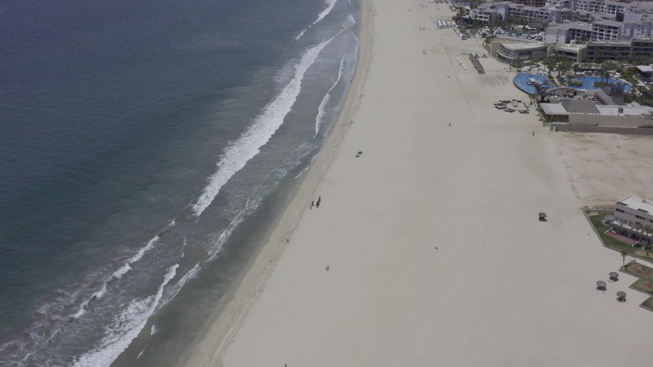 Aerial view of surf rolling beside a wide sandy beach and beachfront buildings.