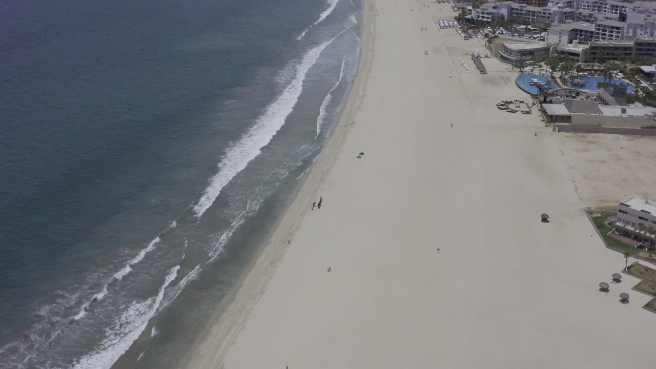 The Los Cabos aerial view holds on white surf crossing the beach edge, open pale sand, small people near the water, and resort-style buildings at the upper right.