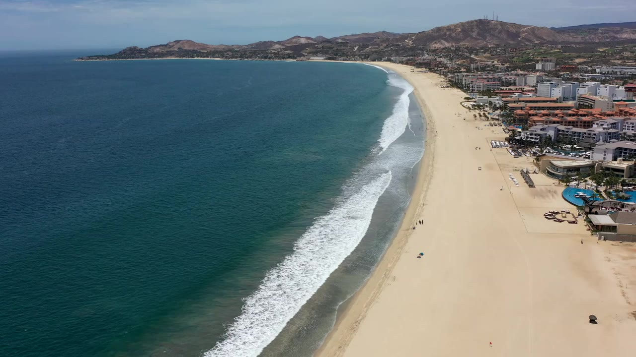 Wide aerial view over a Los Cabos beach with blue ocean on the left, a long white surf line, beachfront buildings on the right, and dry hills in the distance.