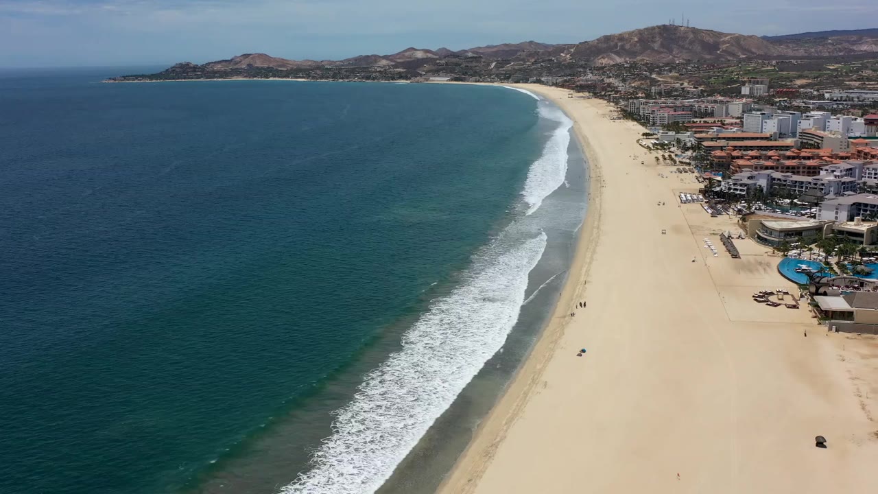 Aerial Los Cabos coastline frame with the ocean filling the left side, waves breaking in a diagonal band, pale sand on the right, and resort buildings beyond the beach.