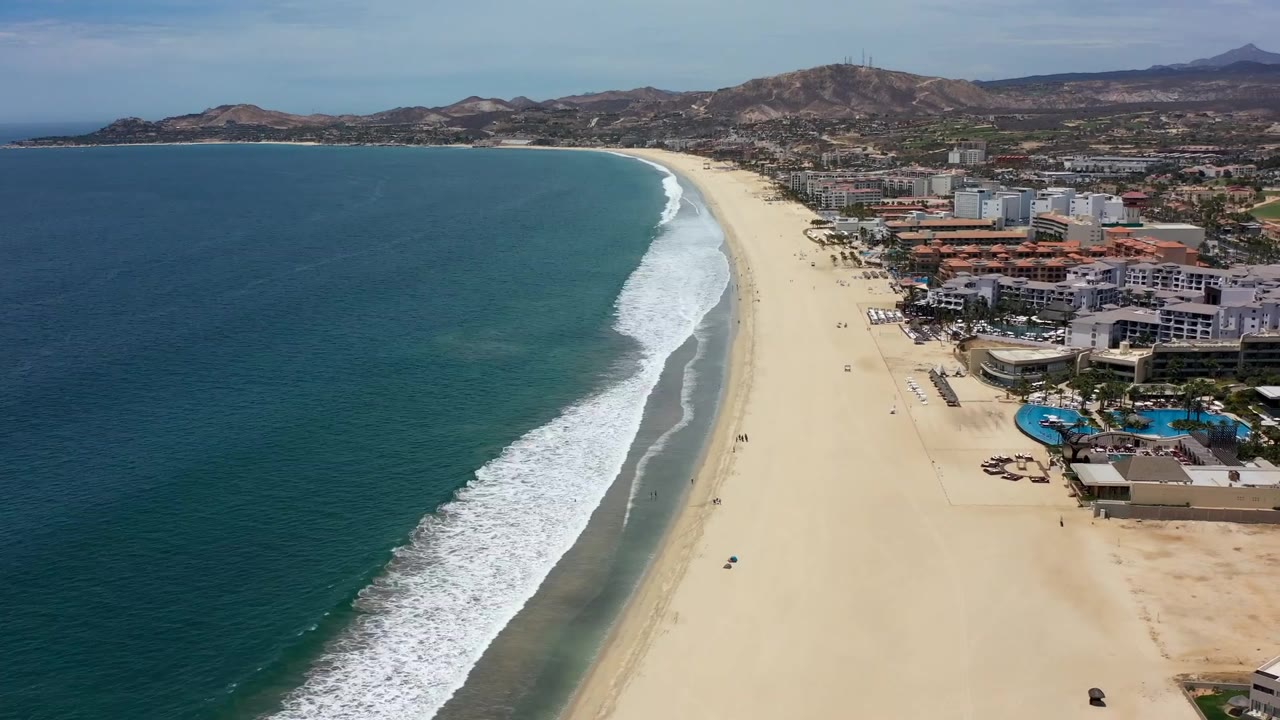 High coastal view of surf, empty sand, beachfront pools, buildings, and hills.