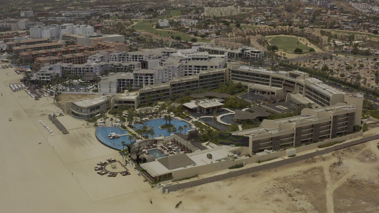 Aerial view of beachfront hotel buildings, pools, sand, and city blocks.