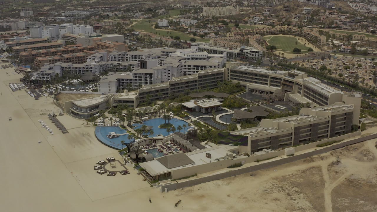 Aerial view in Los Cabos, Mexico with a large hotel complex beside the beach, curved blue pools in the foreground, and urban buildings beyond, with no visible people.