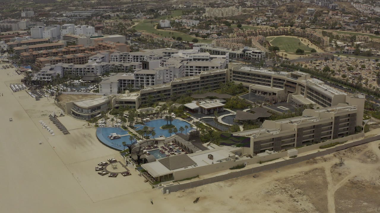 High aerial frame over Los Cabos, Mexico, showing resort buildings, blue pool areas, empty sandy foreground, a main road, distant apartments, and no visible people.