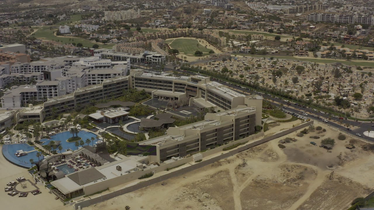 Aerial view in Los Cabos, Mexico showing a U-shaped building complex, blue pools, palm landscaping, roads, sandy open land, and no visible people at the start of the clip.