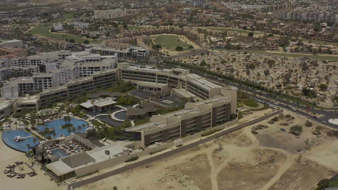 Los Cabos aerial frame with the modern building complex lower in view, curved blue pools to the left, road traffic through the middle distance, and no visible people.