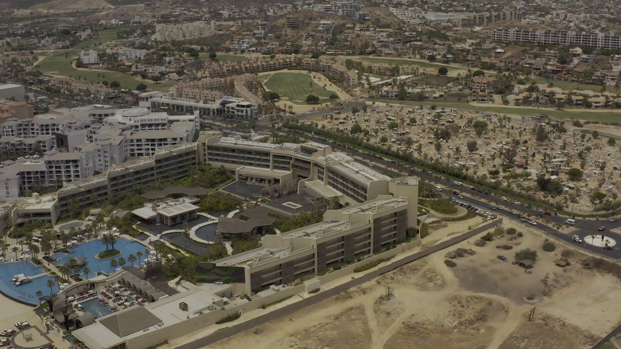 Opening aerial frame over Los Cabos, Mexico, showing a hotel complex, blue pool area, nearby highway, dry open lots, distant buildings, and no visible people.