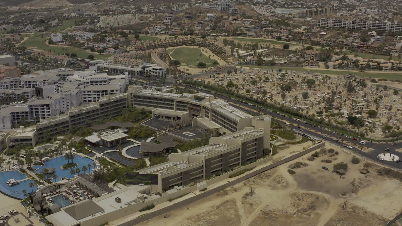 Aerial frame of hotel blocks, a pool, road traffic, open desert-like ground, and city structures.