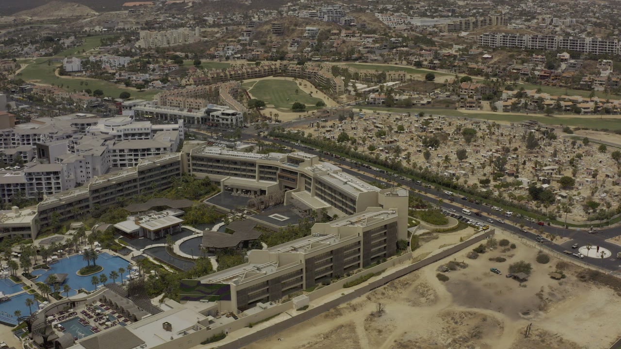 Aerial frame from Los Cabos, Mexico showing resort buildings and blue pools in the foreground, a road with small vehicles through the middle, desert landscaping and distant hills beyond, with no visible people.