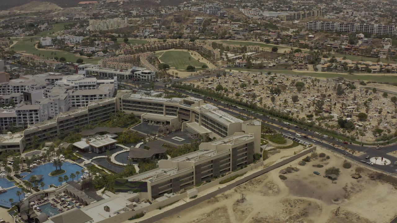 Los Cabos aerial view with tan hotel-style buildings and pool decks at lower left, a divided road crossing the center, sandy undeveloped ground at right, and neighborhoods and hills in the distance, with no visible people.