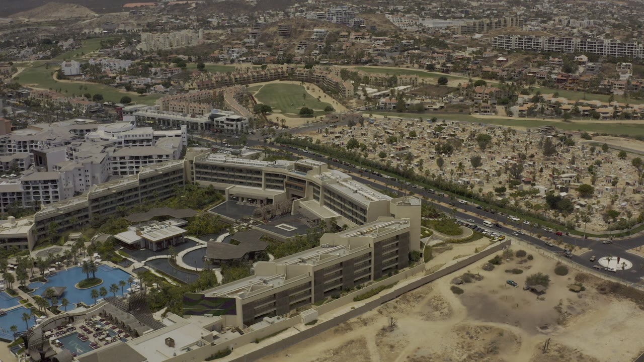Resort complex beside a road with pools, palm trees, desert terrain, and distant residential areas.