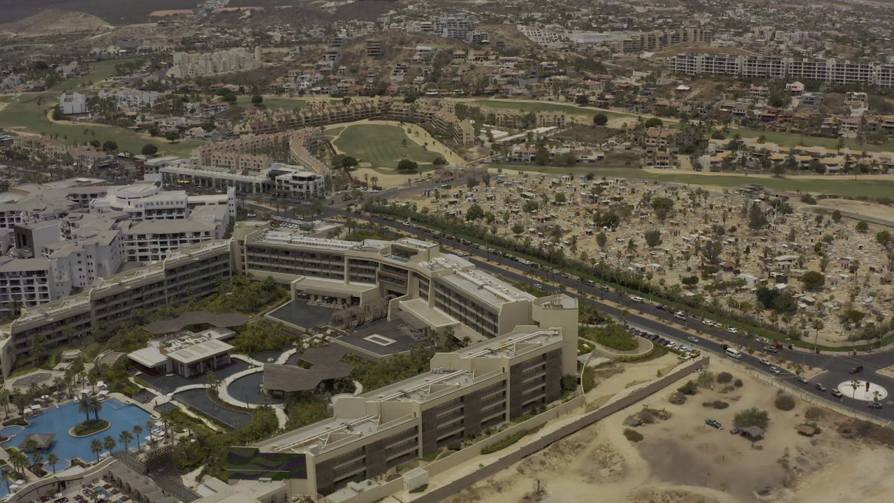 High aerial frame in Los Cabos, Mexico showing resort roofs and landscaped grounds below, a road with small cars beyond them, dry terrain, a green sports field, and distant buildings.