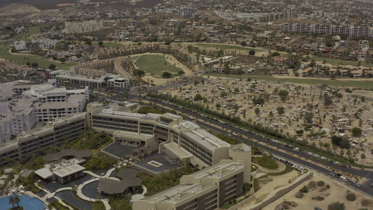 Aerial frame over Los Cabos, Mexico, with large pale buildings in the foreground, a road across the middle, a tree-filled open area, distant neighborhoods, dry hills, and no visible people.
