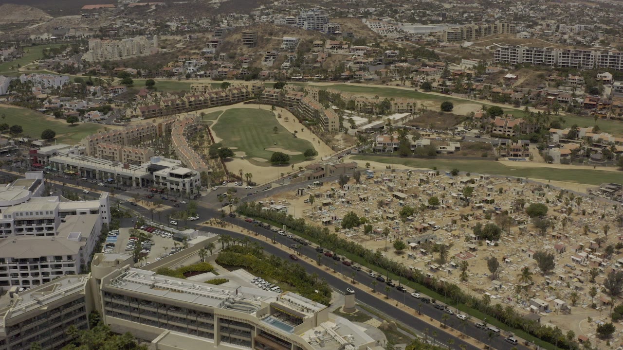 Opening aerial frame over Los Cabos, Mexico, showing roads, low-rise buildings, a cemetery-like area, a green sports field, dry hills, and no visible people.