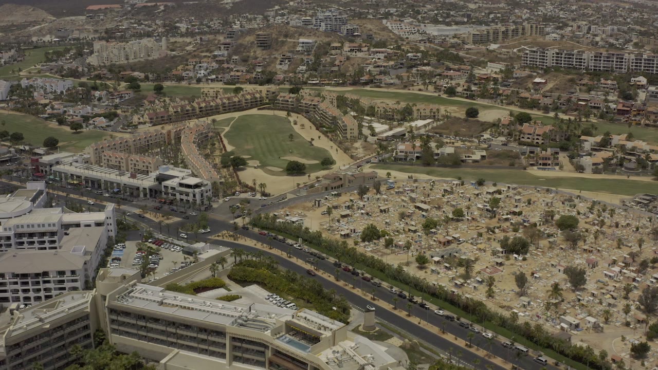 Aerial view in Los Cabos, Mexico, with the cemetery-like grounds centered below, roads crossing the lower frame, housing beyond, and no visible people.