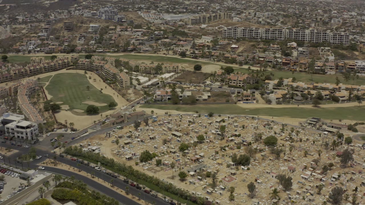 Aerial view in Los Cabos with the dry marker area lower in frame, a road junction at left, a green fairway band through the middle, distant hillside residences, and no visible people.