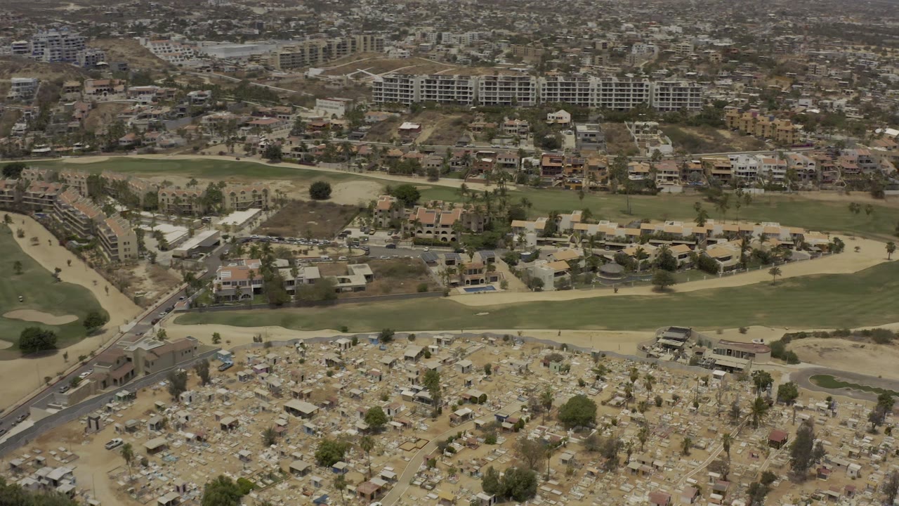 A second aerial moment in Los Cabos holds over dense grave plots at the bottom, a green fairway band across the center, clustered houses beyond it, and no visible people.