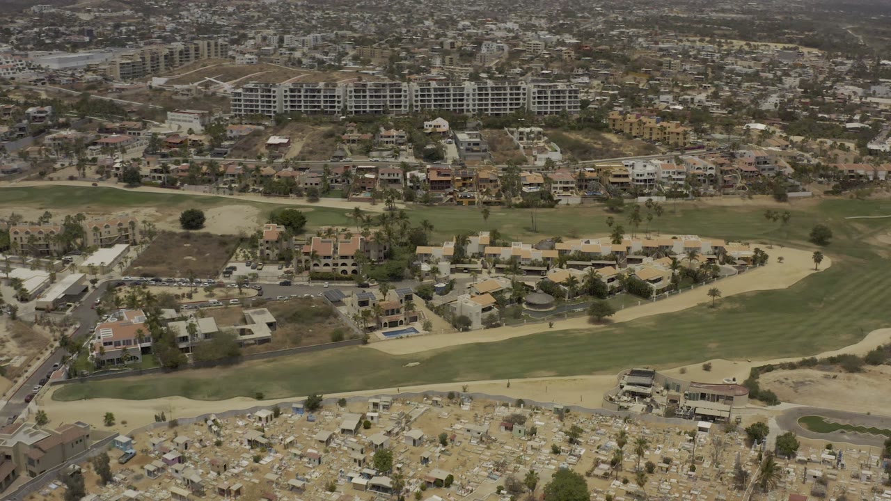 High aerial frame in Los Cabos with striped green grass in the center, homes below and across the midground, larger buildings behind, and no visible people.