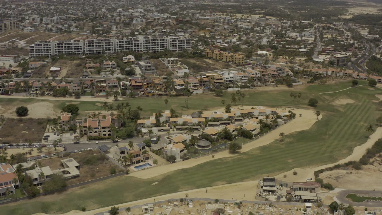 Opening aerial frame over Los Cabos, Mexico, with a striped golf fairway, pale sand bunkers, low houses, roads, hillside apartment buildings, and no visible people.