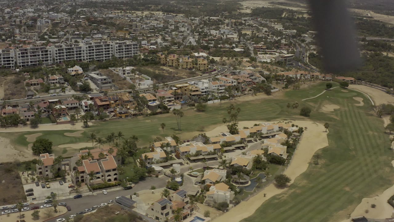 Los Cabos aerial frame with residential homes in the lower frame, a striped green fairway to the right, apartment buildings across the upper frame, and no visible people.
