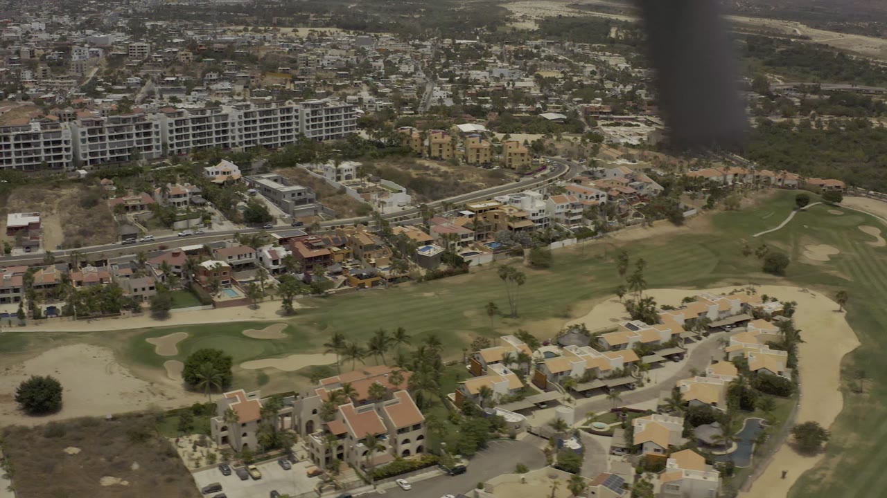 Aerial frame of Los Cabos, Mexico, with apartment blocks at upper left, residential streets below, golf fairways across the middle, and no visible people.
