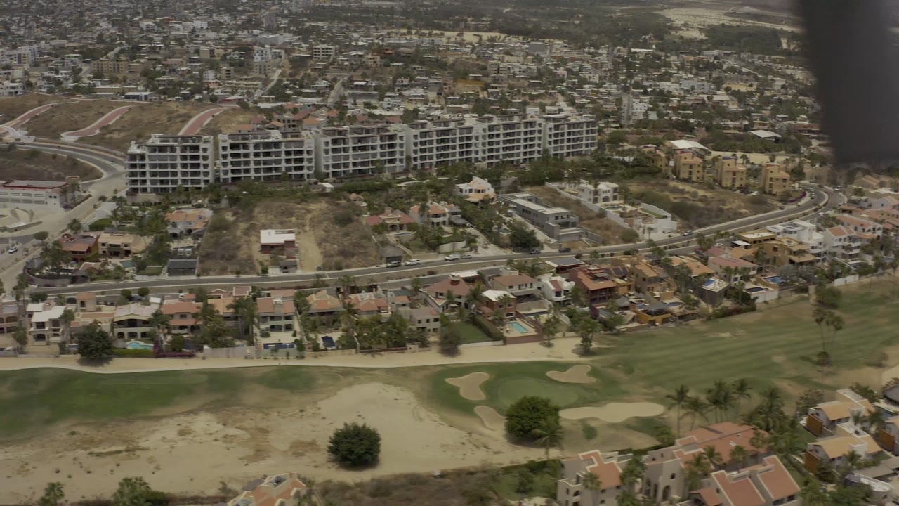 Aerial frame in Los Cabos, Mexico, with green fairways at the bottom, villas and streets in the middle, and a long unfinished building complex on the hillside, with no visible people.
