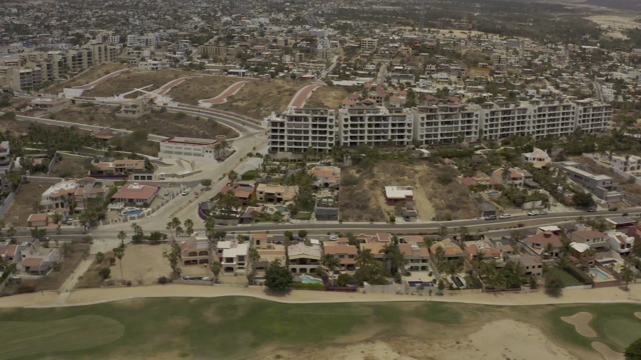 Aerial frame in Los Cabos showing tan open lots, a curved road, clustered homes, larger buildings on the slope, green golf-course turf below, and no visible people.