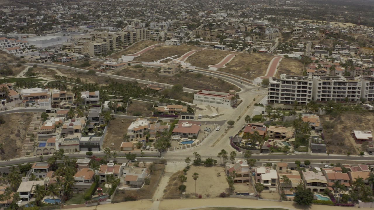 Opening aerial frame over Los Cabos, Mexico showing a residential road, private homes with pools, graded dirt lots, hillside buildings, and no visible people.