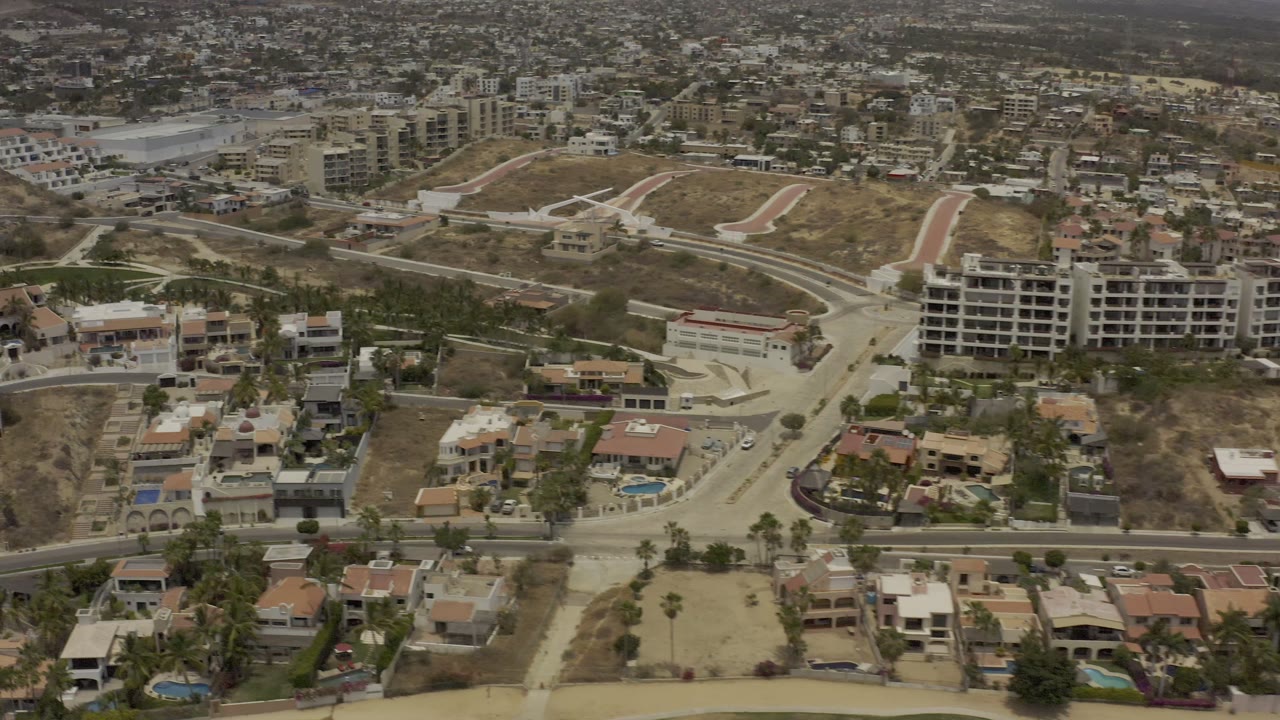 Aerial frame over Los Cabos, Mexico with a central road intersection, tan residential roofs, blue swimming pools, unfinished lots, and no visible people.
