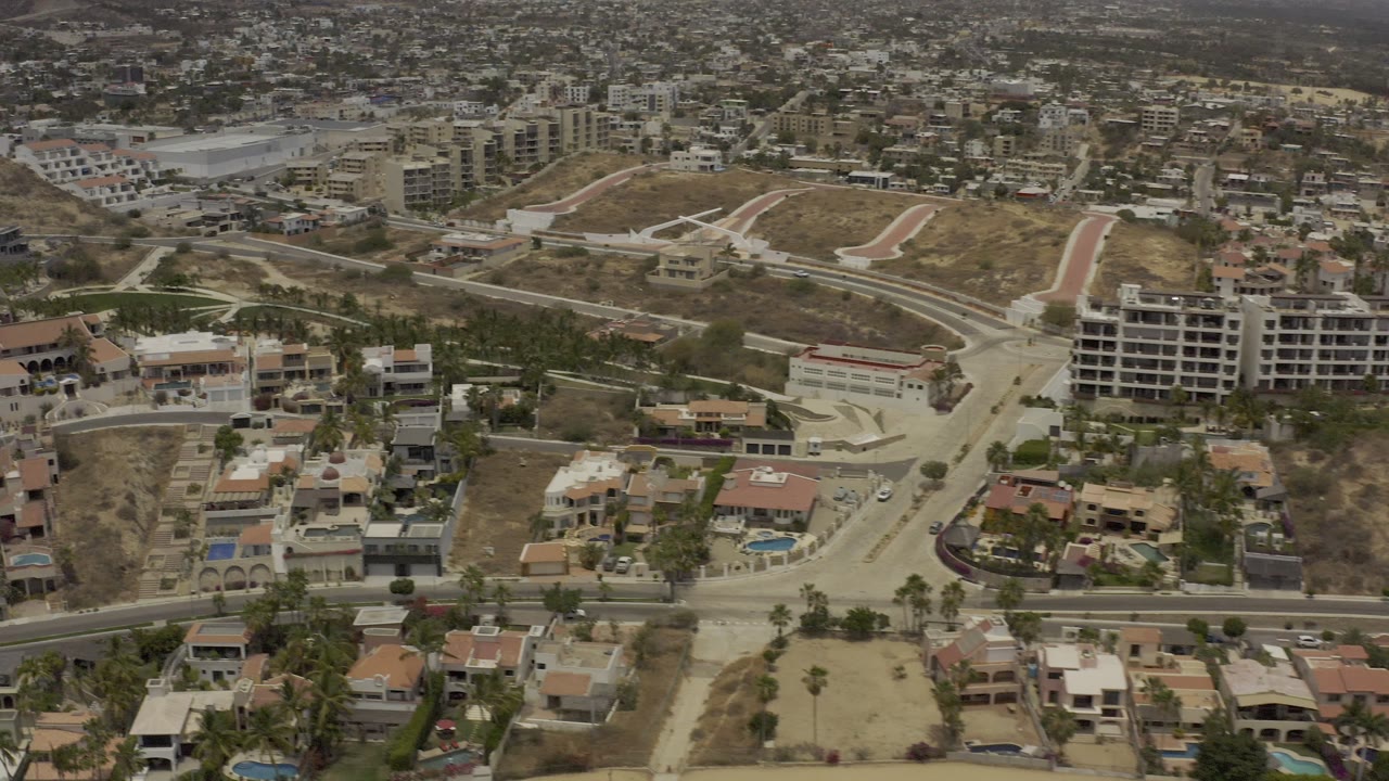 Aerial view across Los Cabos neighborhood streets, pools, palms, and distant city blocks.