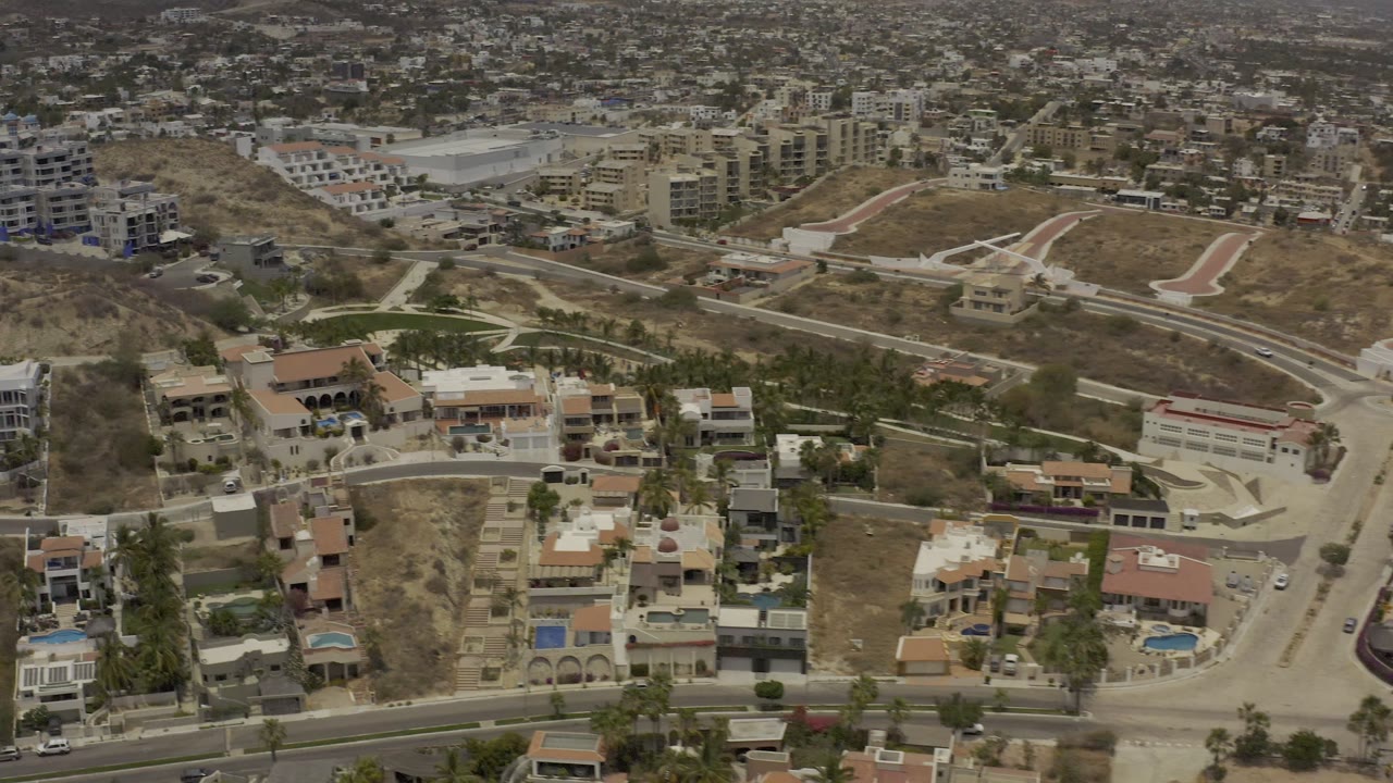 Los Cabos aerial view showing hillside houses, a winding road, open dirt lots, low-rise buildings, and distant city blocks with no visible people.