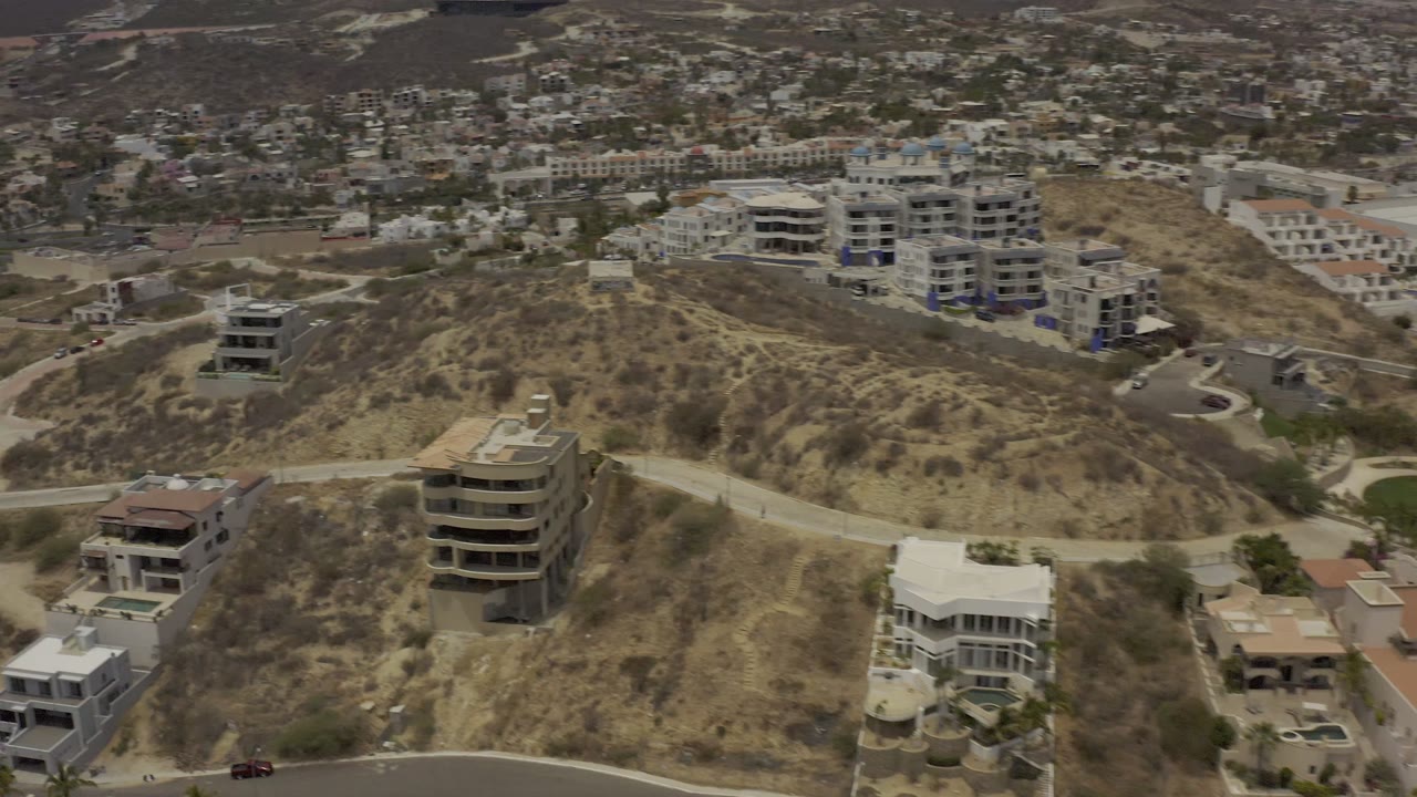 Los Cabos aerial frame showing a pale road across scrubby hills, several modern residences, apartment blocks in the background, and no visible people.