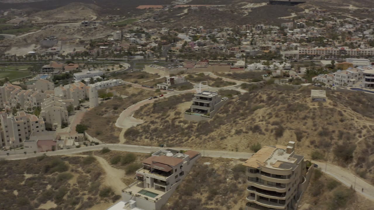 Early aerial view of Los Cabos with pale apartment buildings at left, a curving road through dry hills, scattered homes, distant neighborhoods, and no visible people.