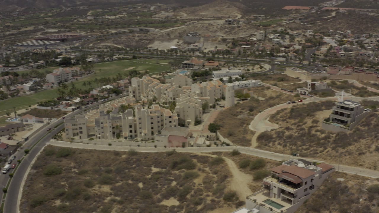 Aerial view over Los Cabos with cream apartment buildings, curving roads, dry hillside lots, green fields, and no visible people.