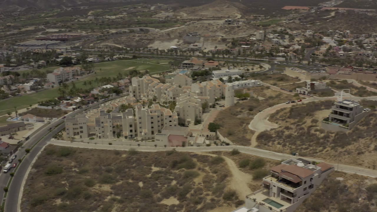 High aerial view in Los Cabos showing apartment blocks beside a curved road, pale dirt paths, scattered houses, green turf, and no visible people.