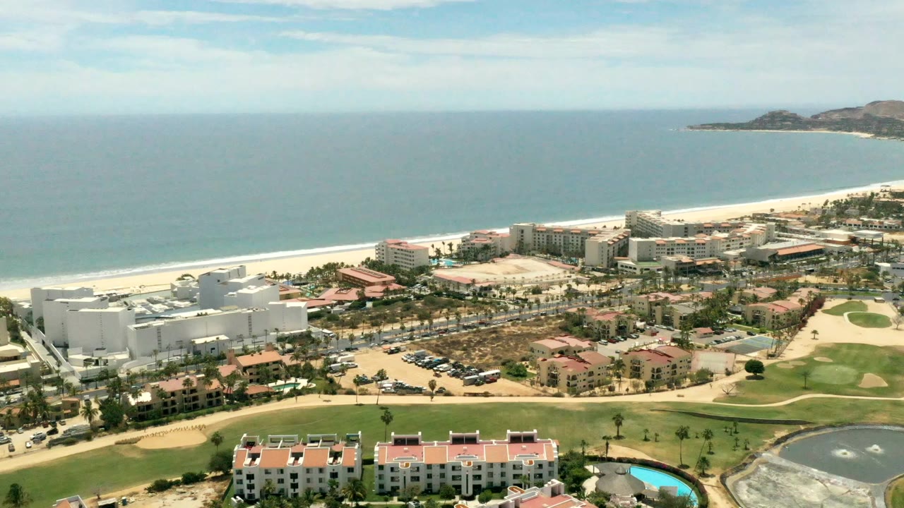 Los Cabos aerial frame showing ocean horizon, pale beach, white and red-roofed beachfront buildings, palm-lined roads, golf course areas, and no visible people.