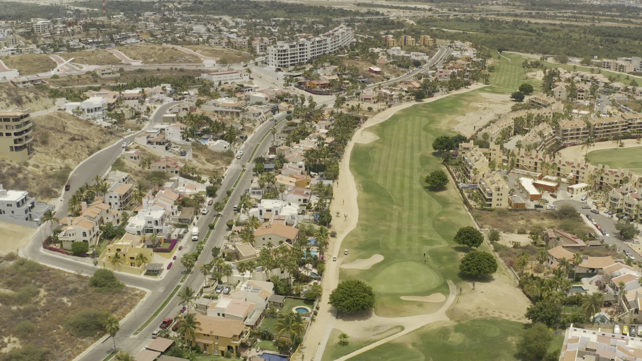 Opening aerial view in Los Cabos, Mexico, showing a green golf fairway, sand traps, curved roads, hillside homes, and distant buildings with no visible people.