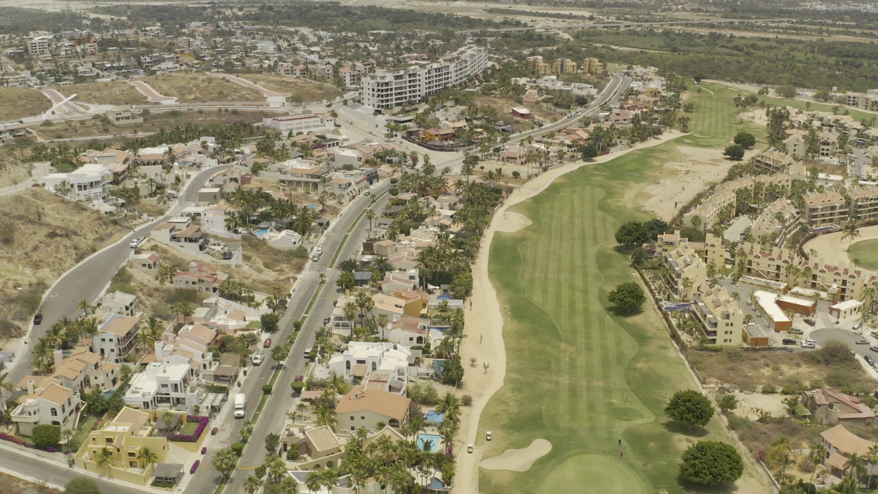 A high aerial frame over Los Cabos shows a long green golf fairway bordered by roads, villas, apartment buildings, palms, and dry hills, with no visible people.
