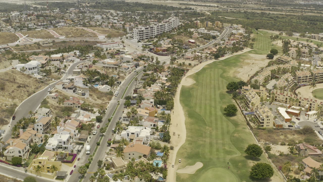 The second frame keeps the Los Cabos fairway centered, with sandy bunkers, hillside homes, white buildings, parked vehicles, and arid land beyond, with no visible people.