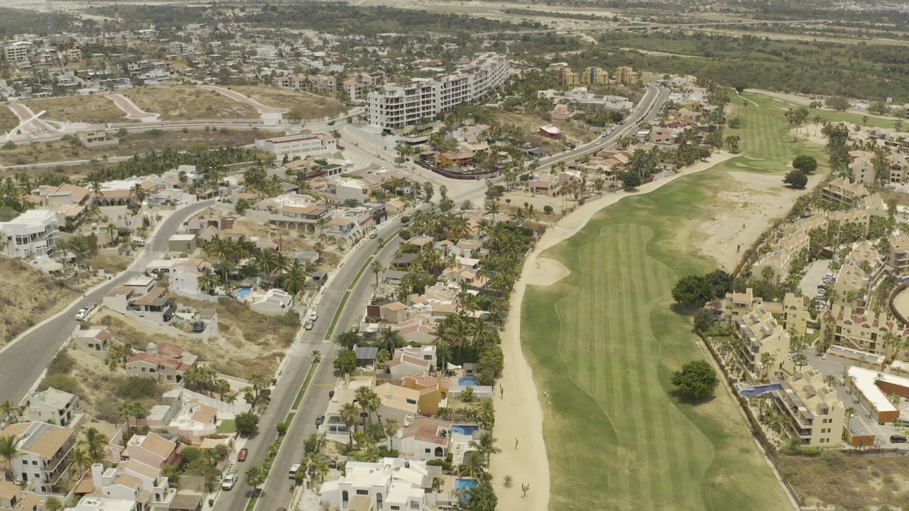 Second aerial frame in Los Cabos, Mexico, with the fairway centered, houses and palms along both sides, dry lots nearby, and no visible people.