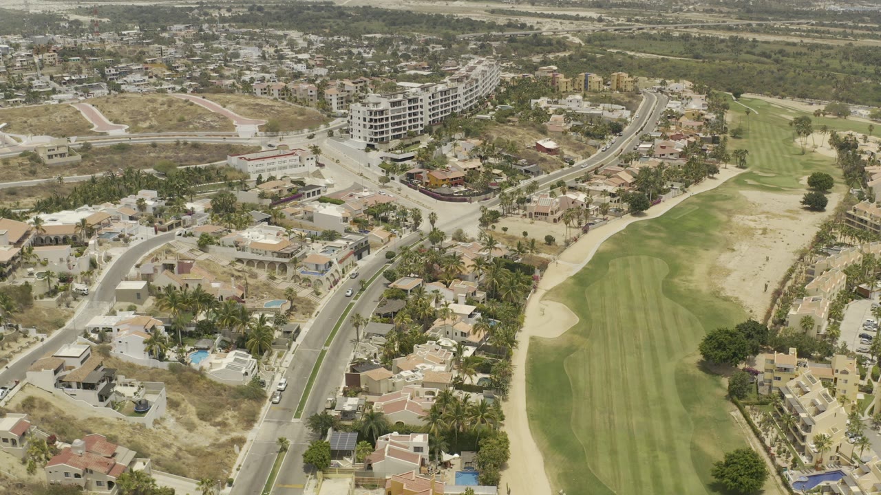 Opening aerial frame over Los Cabos, Mexico with villas, curved streets, palm trees, a green golf fairway, dry lots, and no visible people.