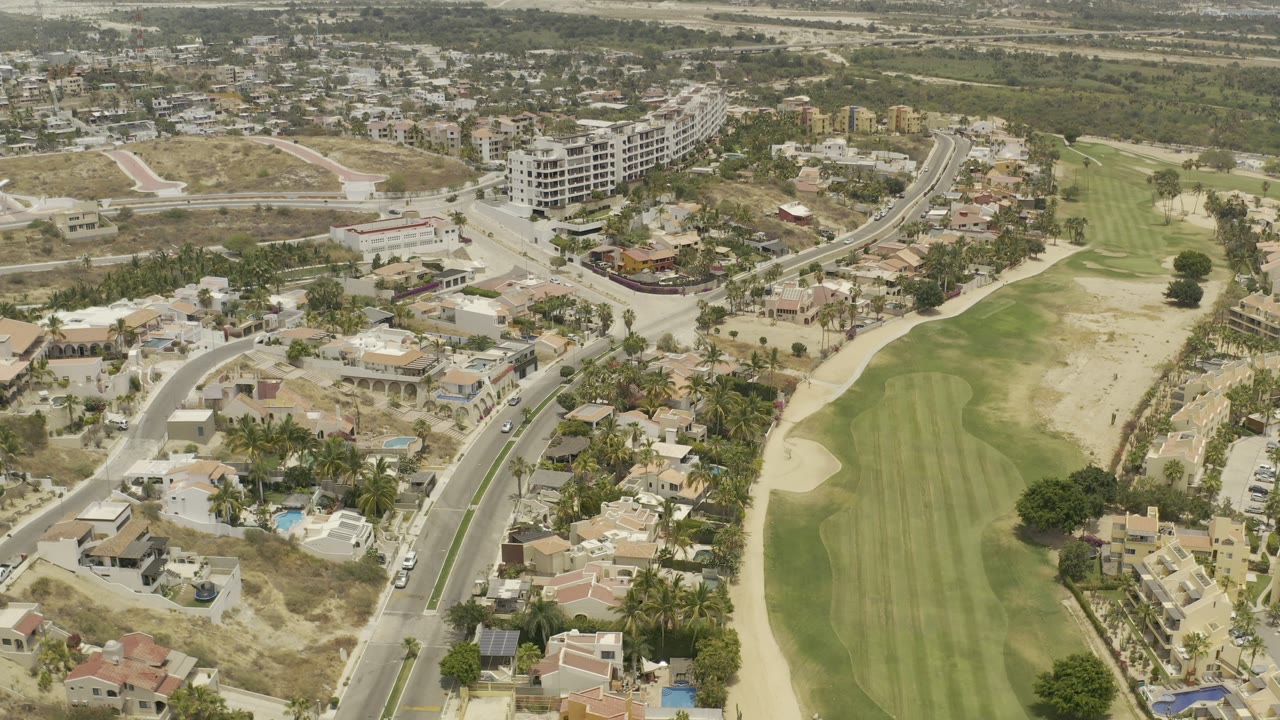 Aerial frame in Los Cabos, Mexico showing pale-roofed villas, a road junction, palm-lined properties, the golf fairway to the right, and no visible people.