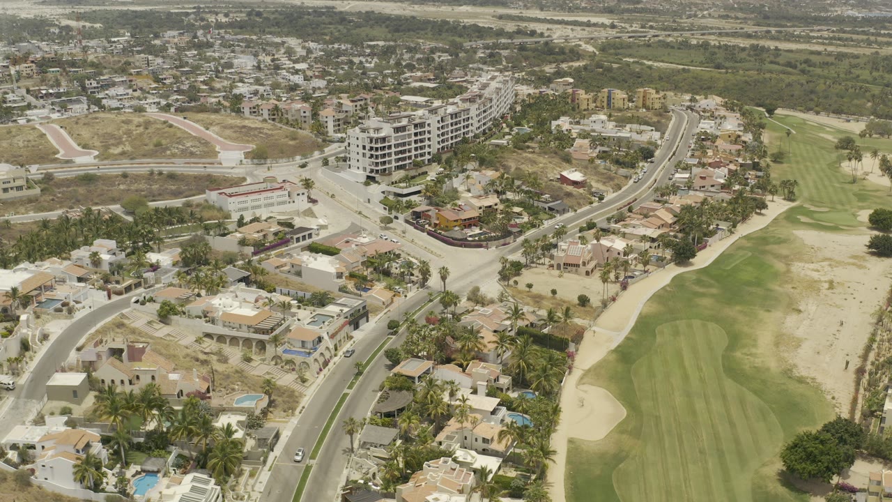 High aerial frame in Los Cabos with a road junction, villas, palm-lined lots, and a golf course running along the right side, with no visible people.