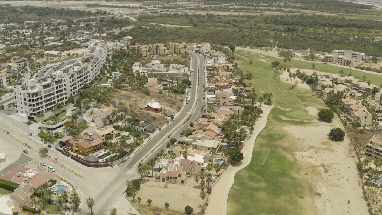 High aerial frame over Los Cabos, Mexico, showing a curved road, white condo building, houses, pools, and a green golf fairway with no visible people.