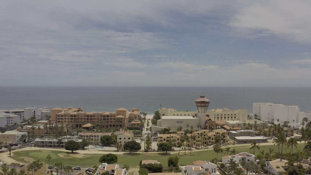 Elevated coastal view with ocean, buildings, palms, streets, and green grounds.