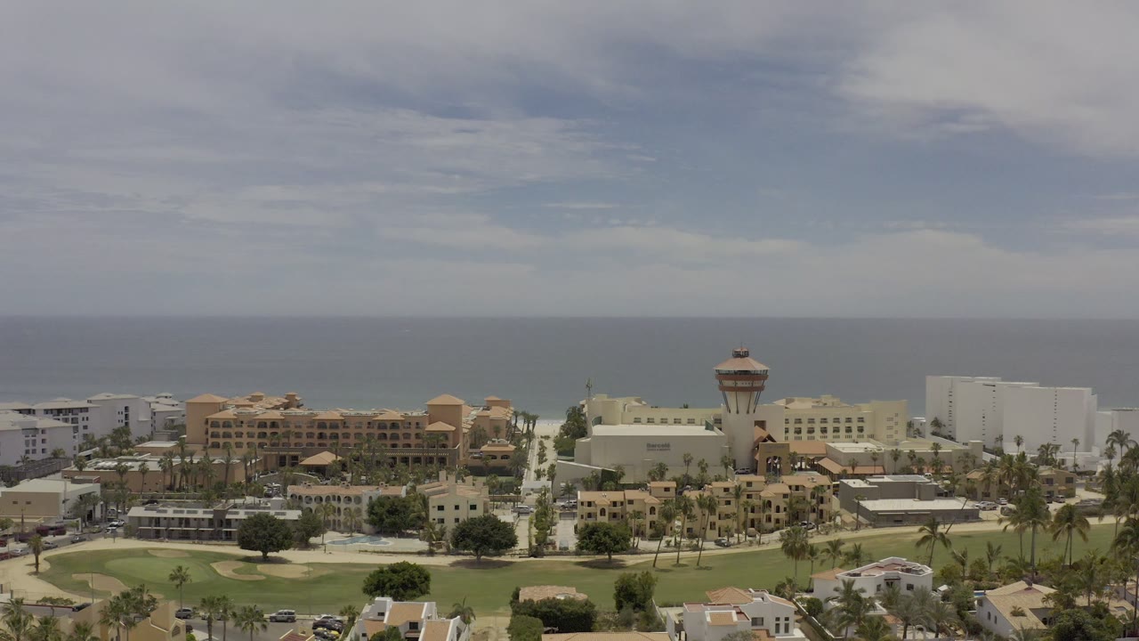 Elevated Los Cabos frame with the tower structure near center, resort-style buildings, white blocks, palms, cars, green grounds, and no visible people.