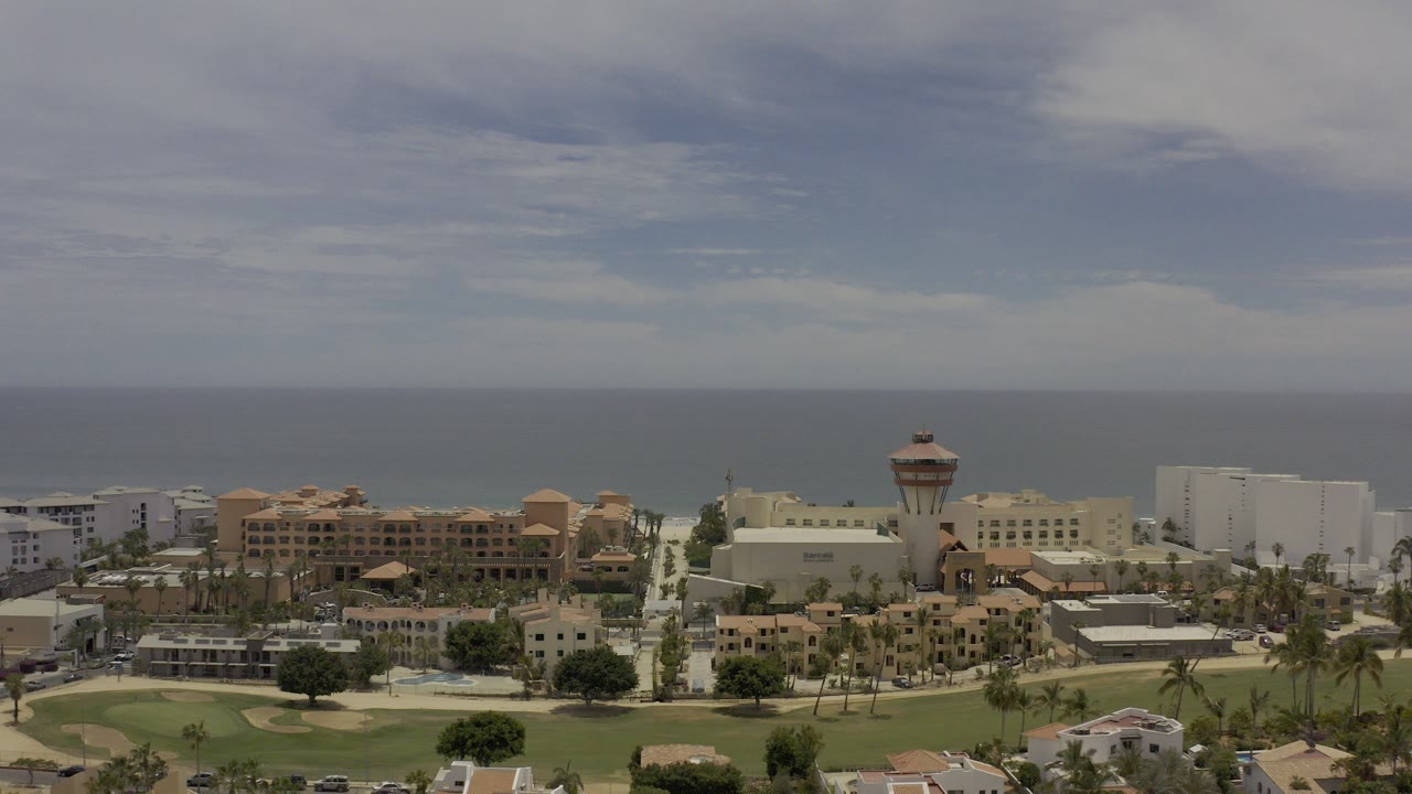 Wide aerial view over Los Cabos coastal buildings and palms toward the ocean horizon, with no visible people discernible in the frame.