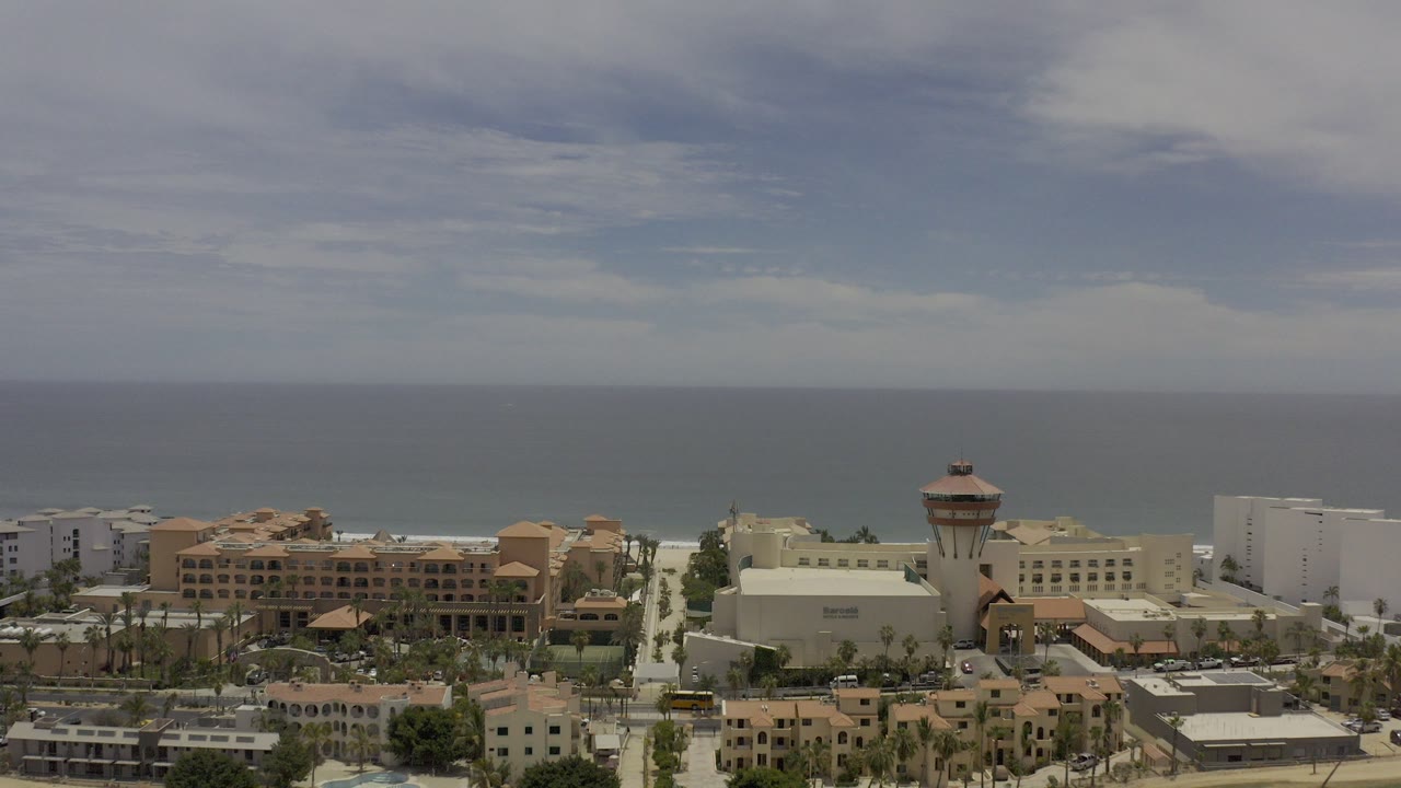 Opening aerial frame over Los Cabos with tan and white buildings, a tower structure, palm-lined streets, beach, ocean horizon, and no visible people.